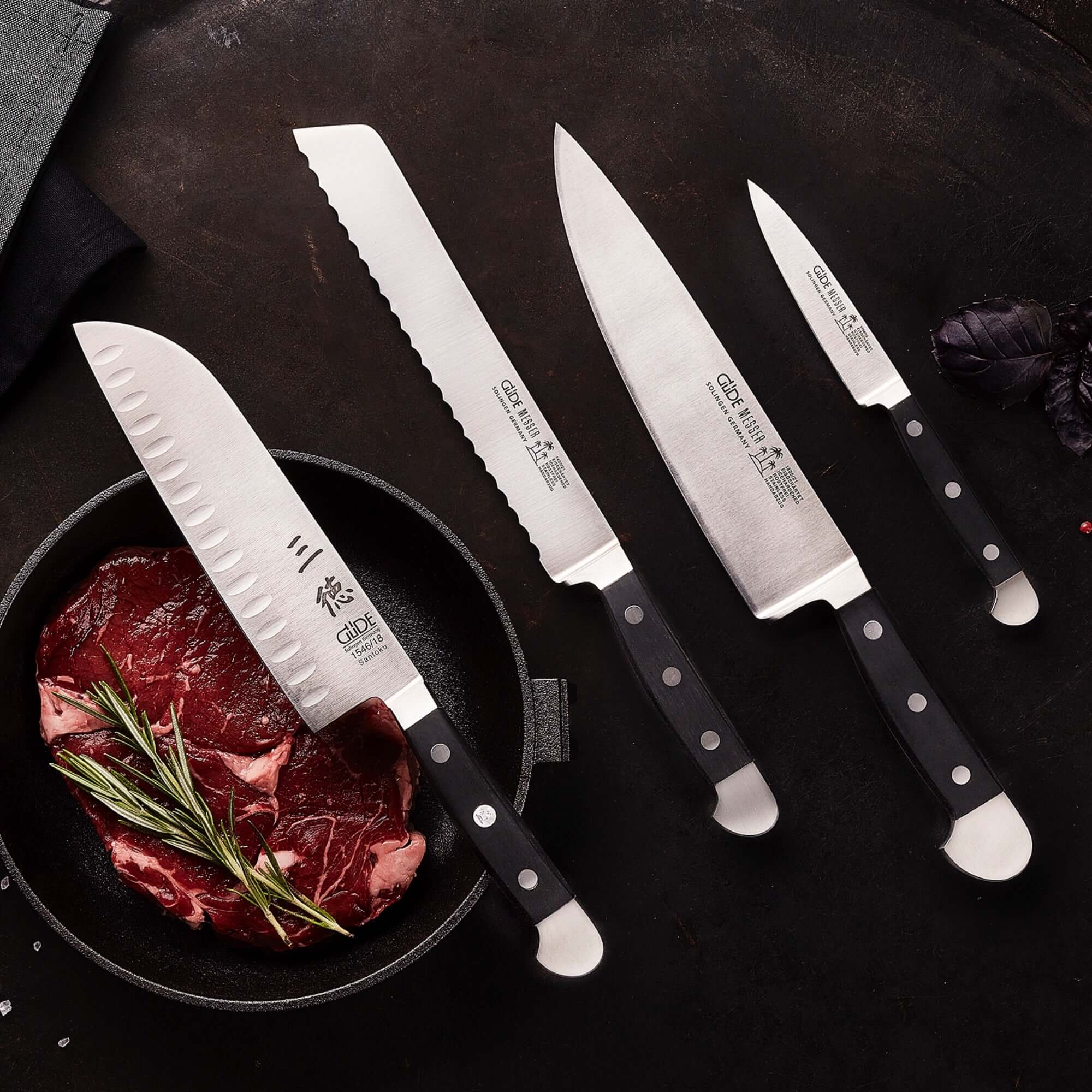 GÜDE | Alpha Series Knives displayed with a variety of blades next to raw meat and herbs.