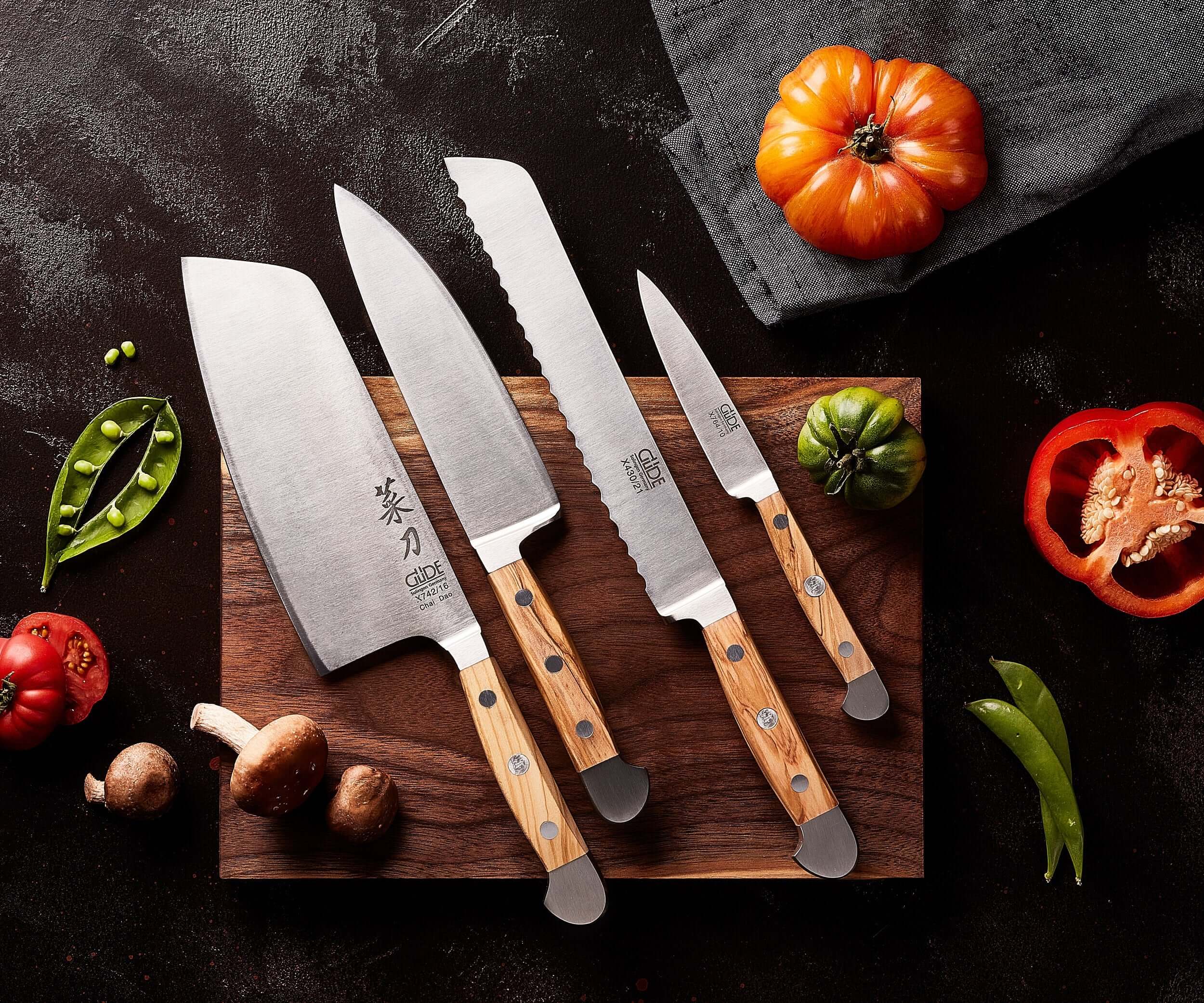 GÜDE | ALPHA OLIVE Series chef knives displayed on a wooden cutting board with fresh vegetables.