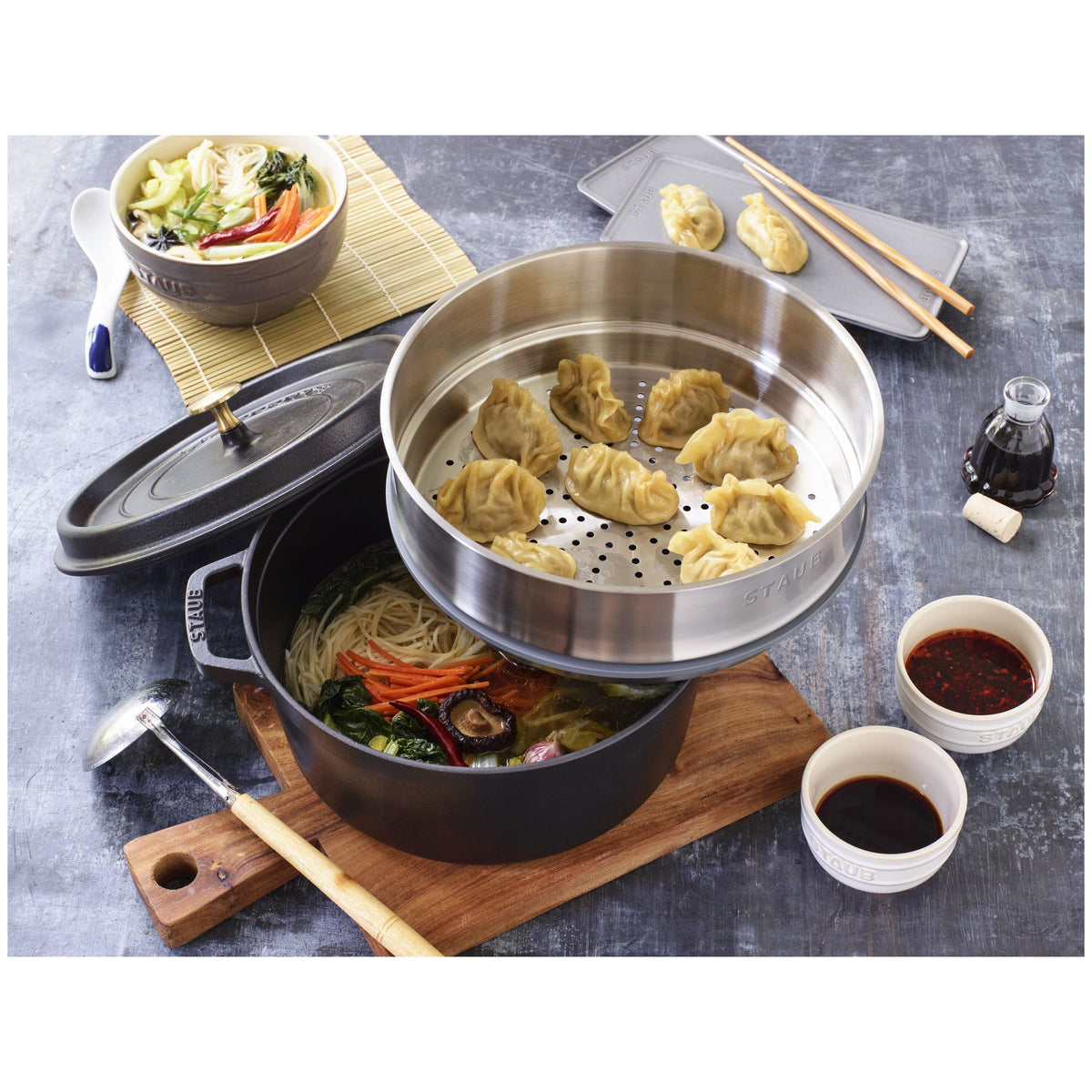 Steaming pot with dumplings and vegetables on a table with condiments and chopsticks.