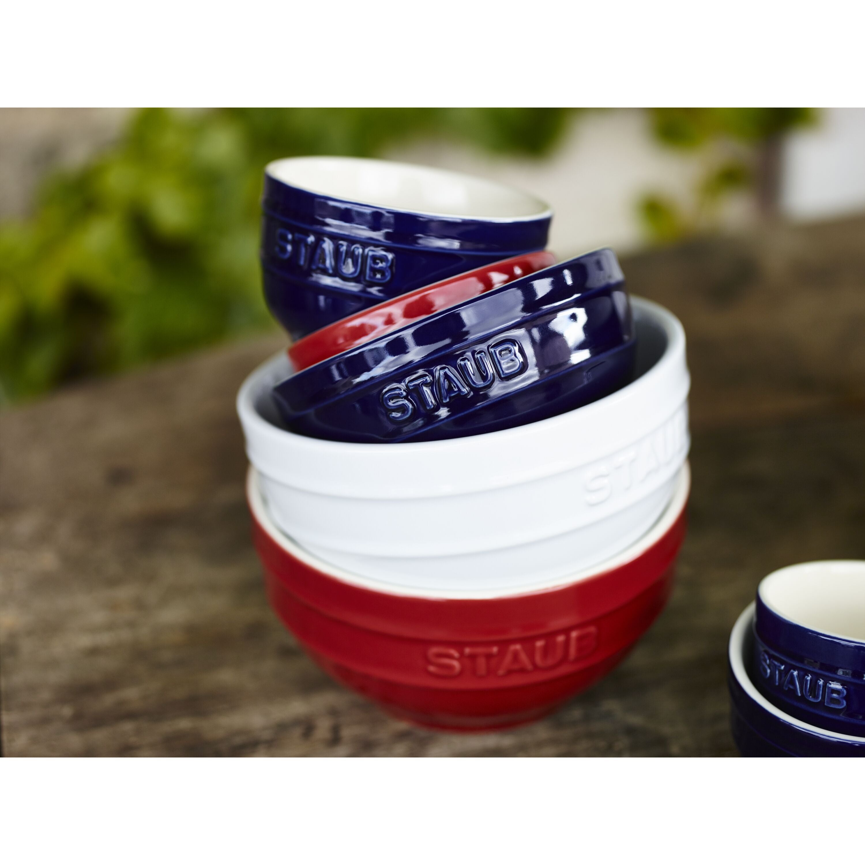 Stacked STAUB Ceramique Ceramic Round Bowls in red, blue, and white on wooden surface with greenery in background.