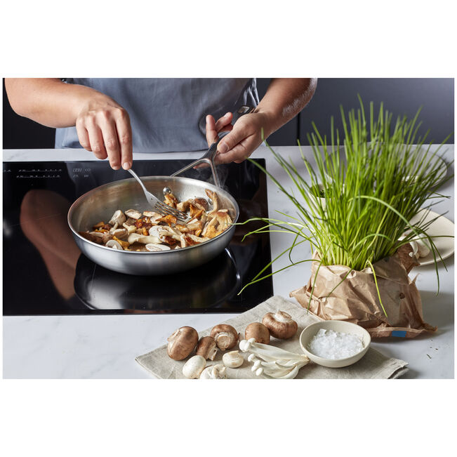 Person cooking mushrooms in a pan on a stove with ingredients and plants on the side.