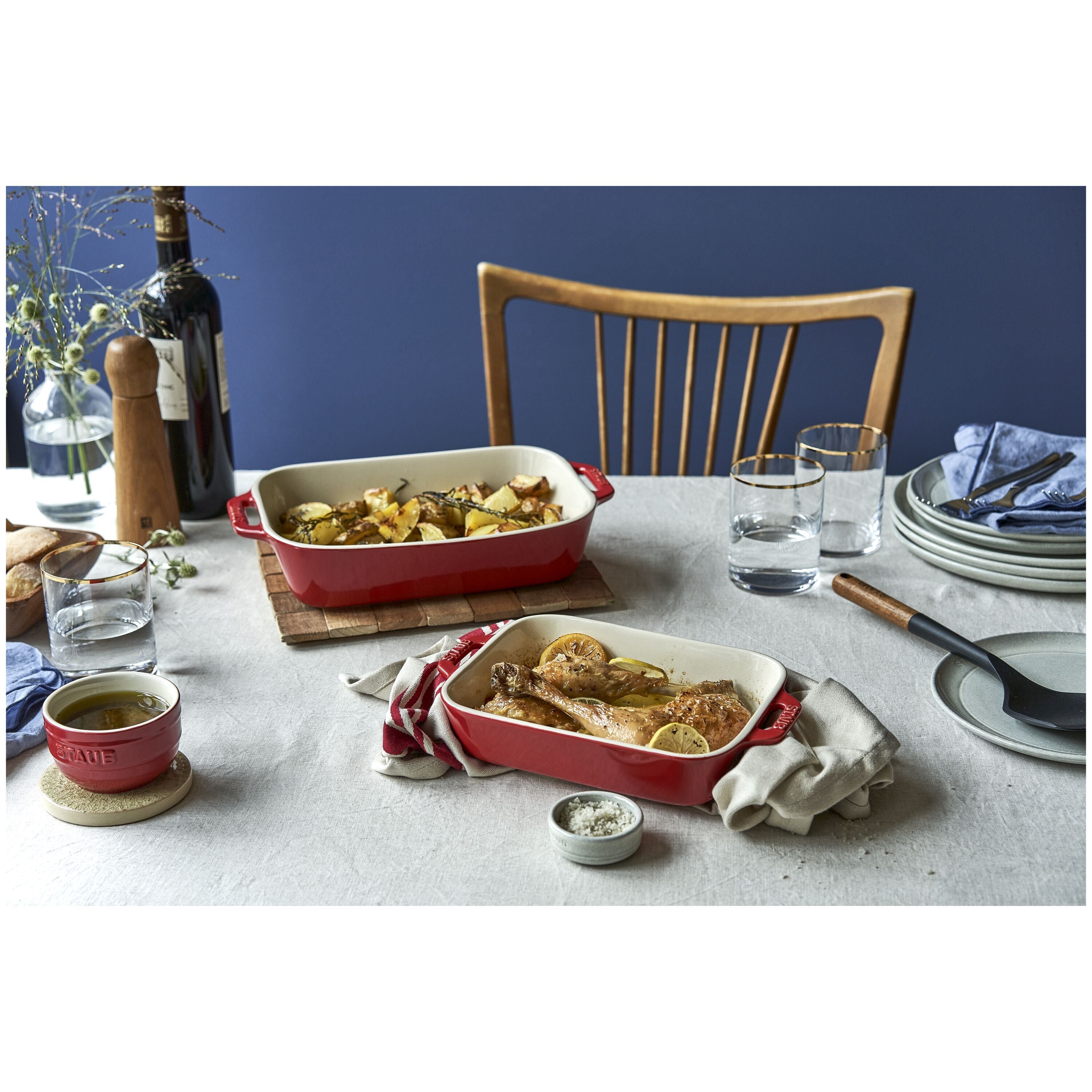 Dining table set with red STAUB Ceramique Ceramic Rectangular Oven Dish containing roasted meats and vegetables, glasses, and a bottle of wine.