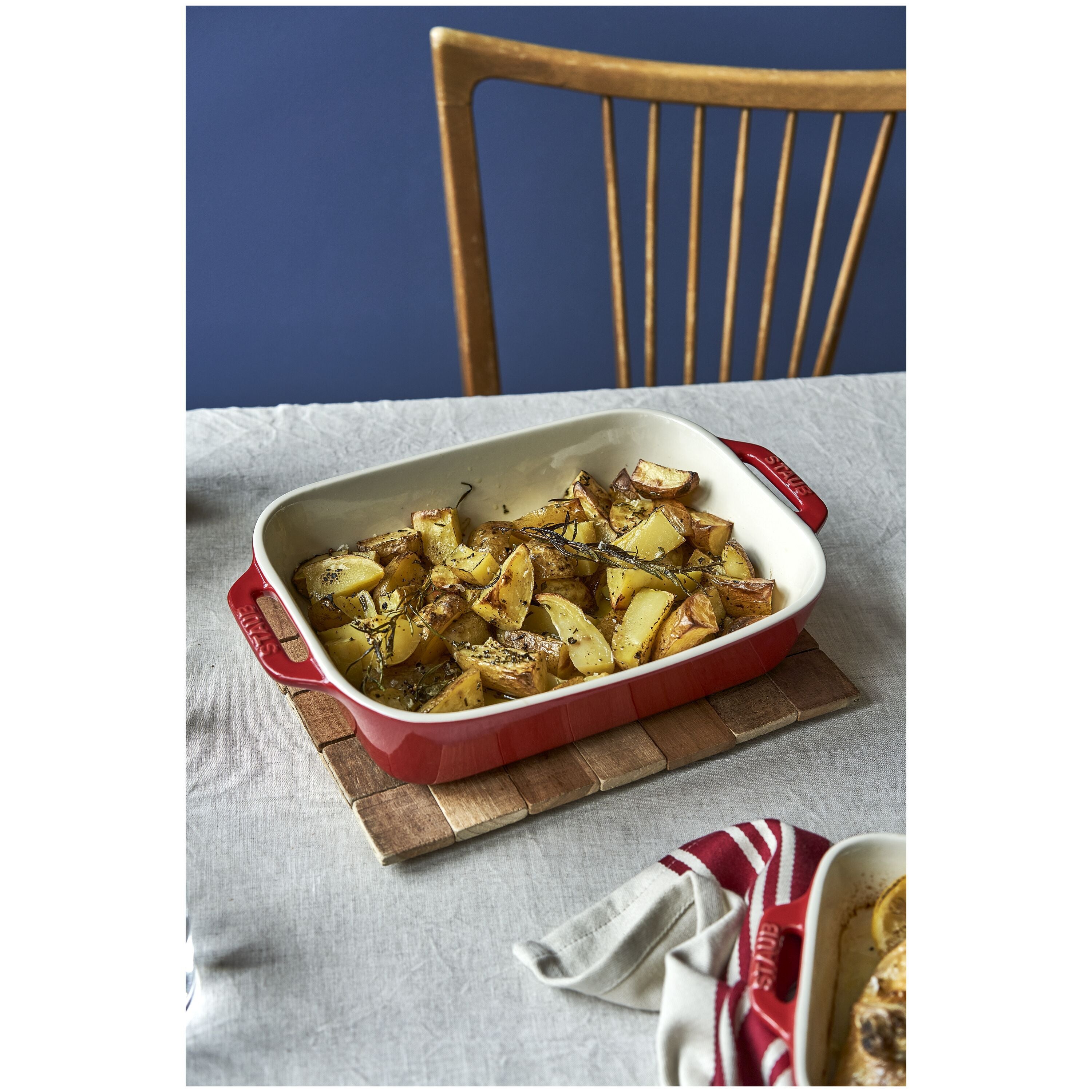 STAUB Ceramique Ceramic Rectangular Oven Dish with roasted potatoes on a wooden board on a table with a blue wall and wooden chair in the background.