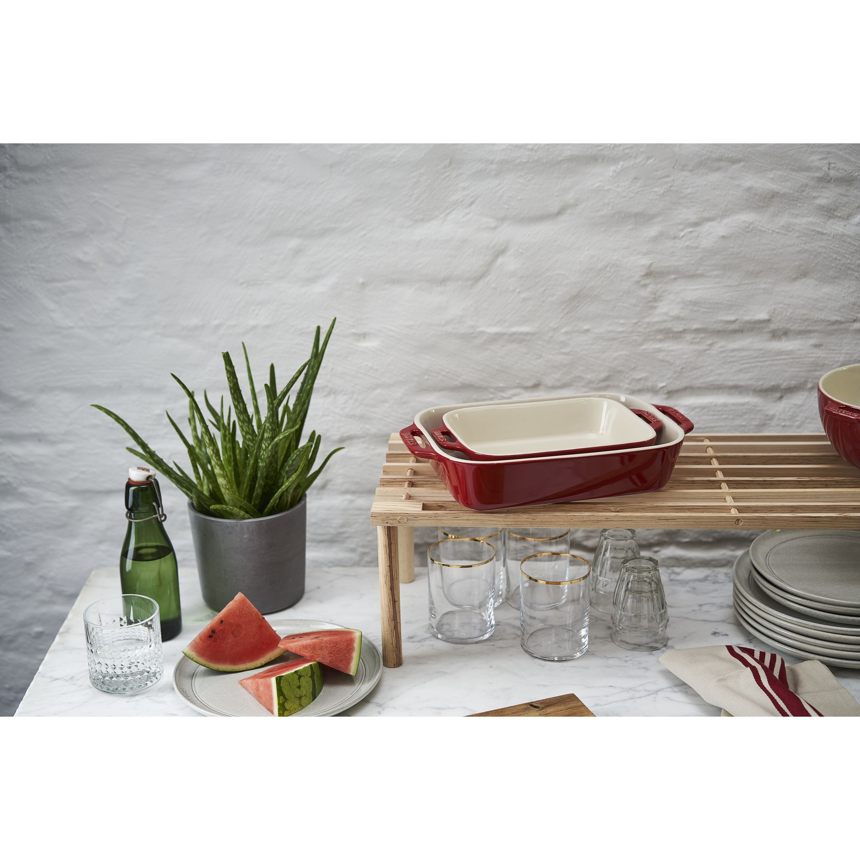 Red ceramic baking dish on a wooden stand with a plant, watermelon, and glasses on a white surface.