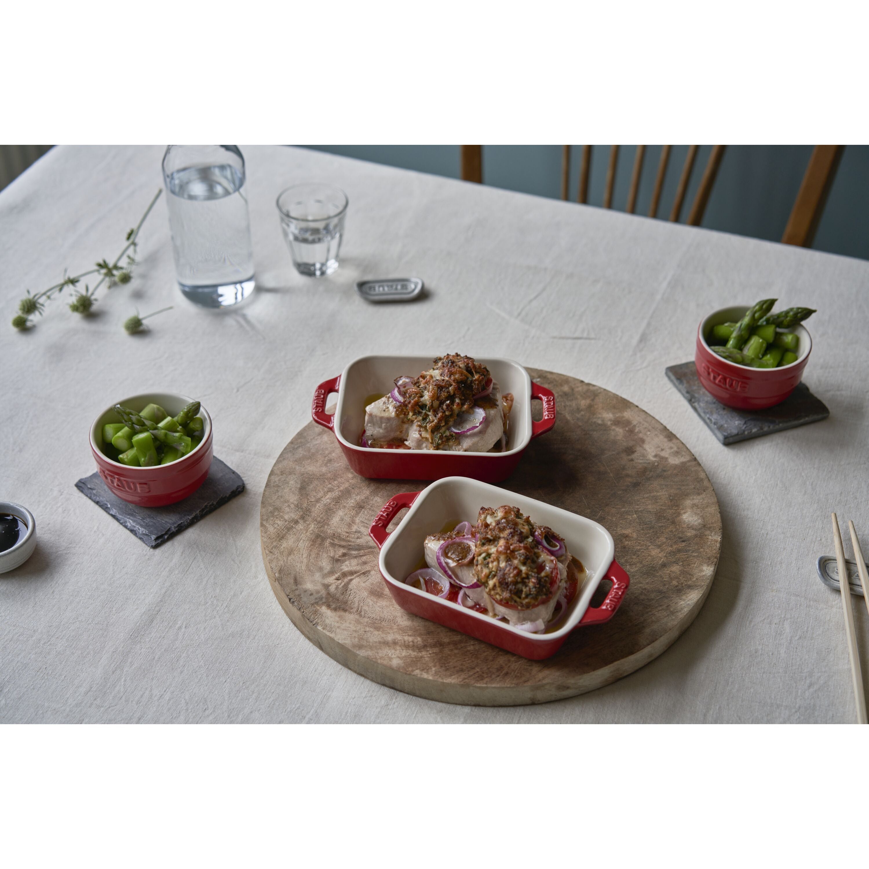 Dinner table setting with red STAUB Ceramique Ceramic Rectangular Oven Dish containing food on a wooden board.