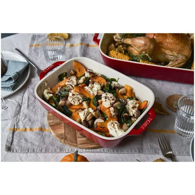 Red casserole dish with roasted vegetables on a table setting with glasses and plates.