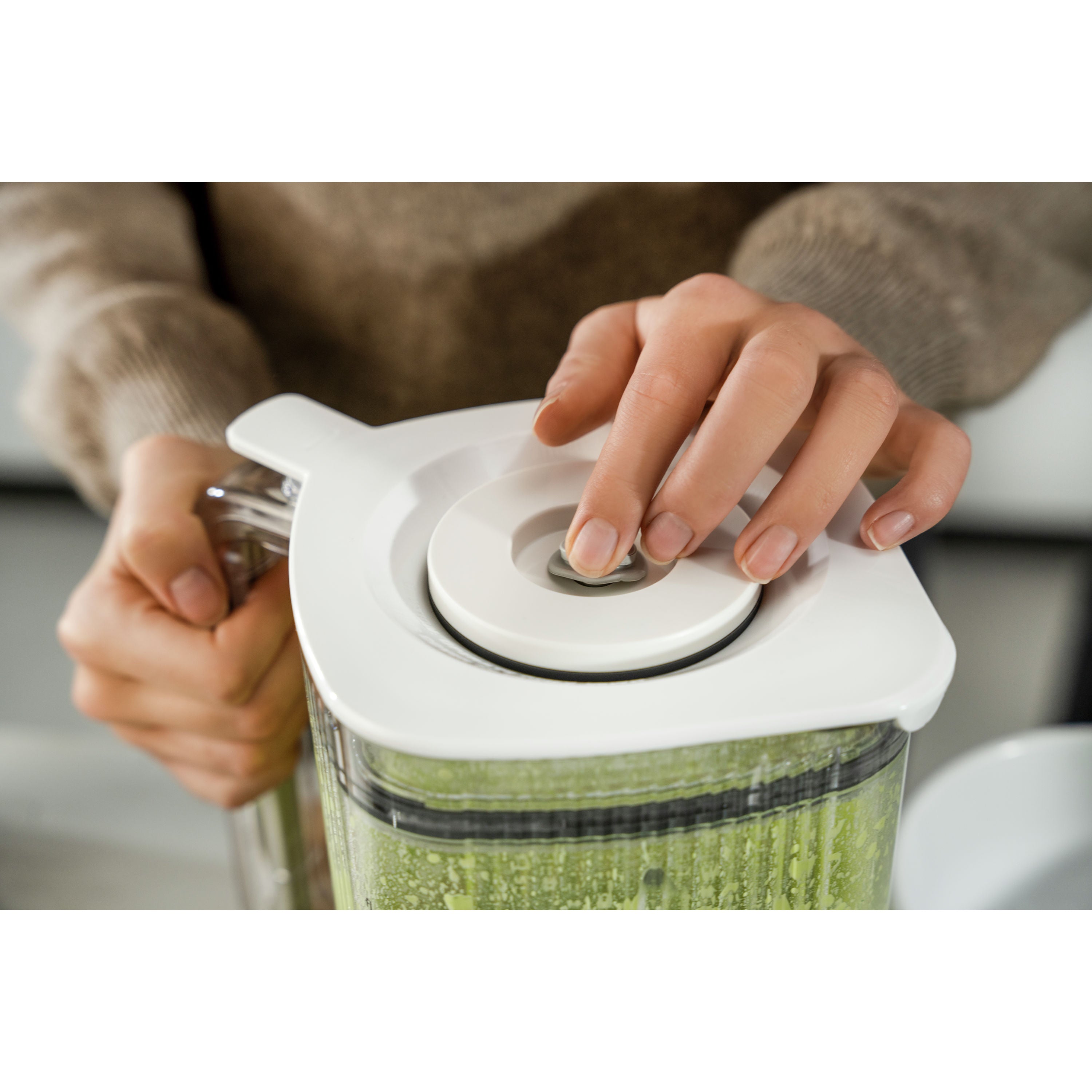 Person using a blender with a green smoothie inside, wearing a brown sweater.