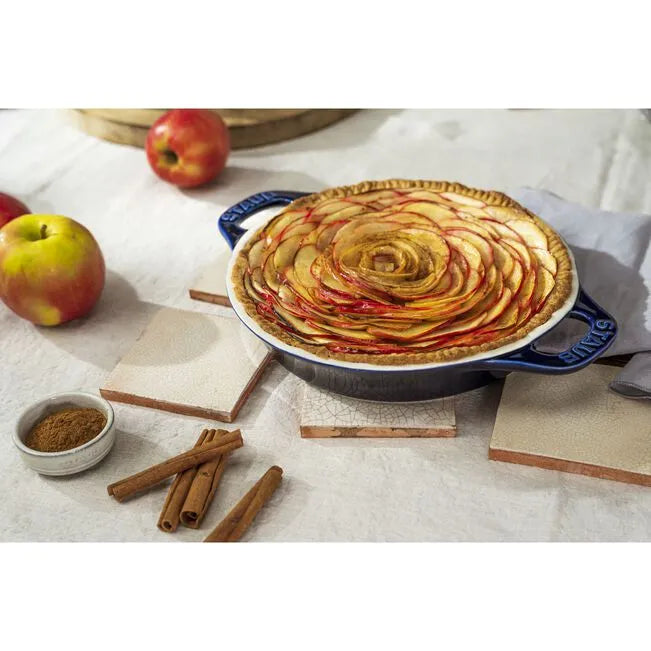 Apple pie with a decorative top in STAUB Ceramique Ceramic Round Pie Dish, surrounded by apples and cinnamon sticks on a white surface.