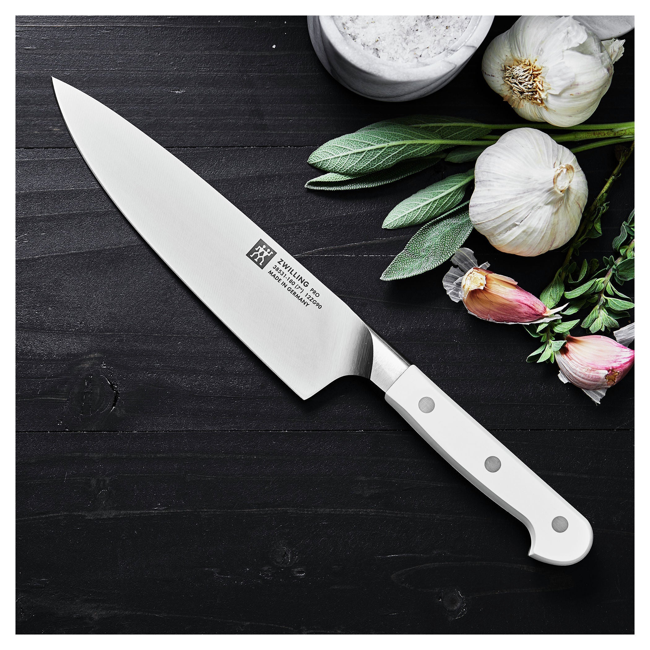 ZWILLING Pro le blanc 7 inch Chef's Knife,with a brand logo on a dark surface with garlic and herbs.