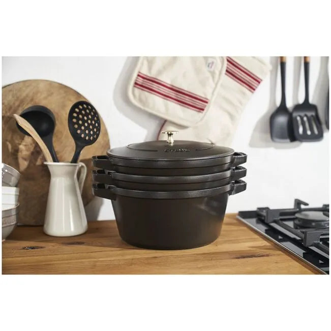 Set of black cookware on a kitchen counter with utensils and a towel in the background.