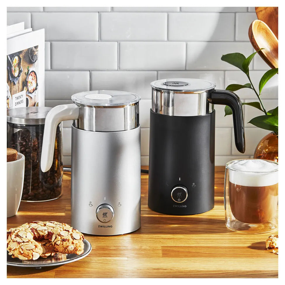 Two milk frothers on a kitchen counter with a glass of coffee and cookies.