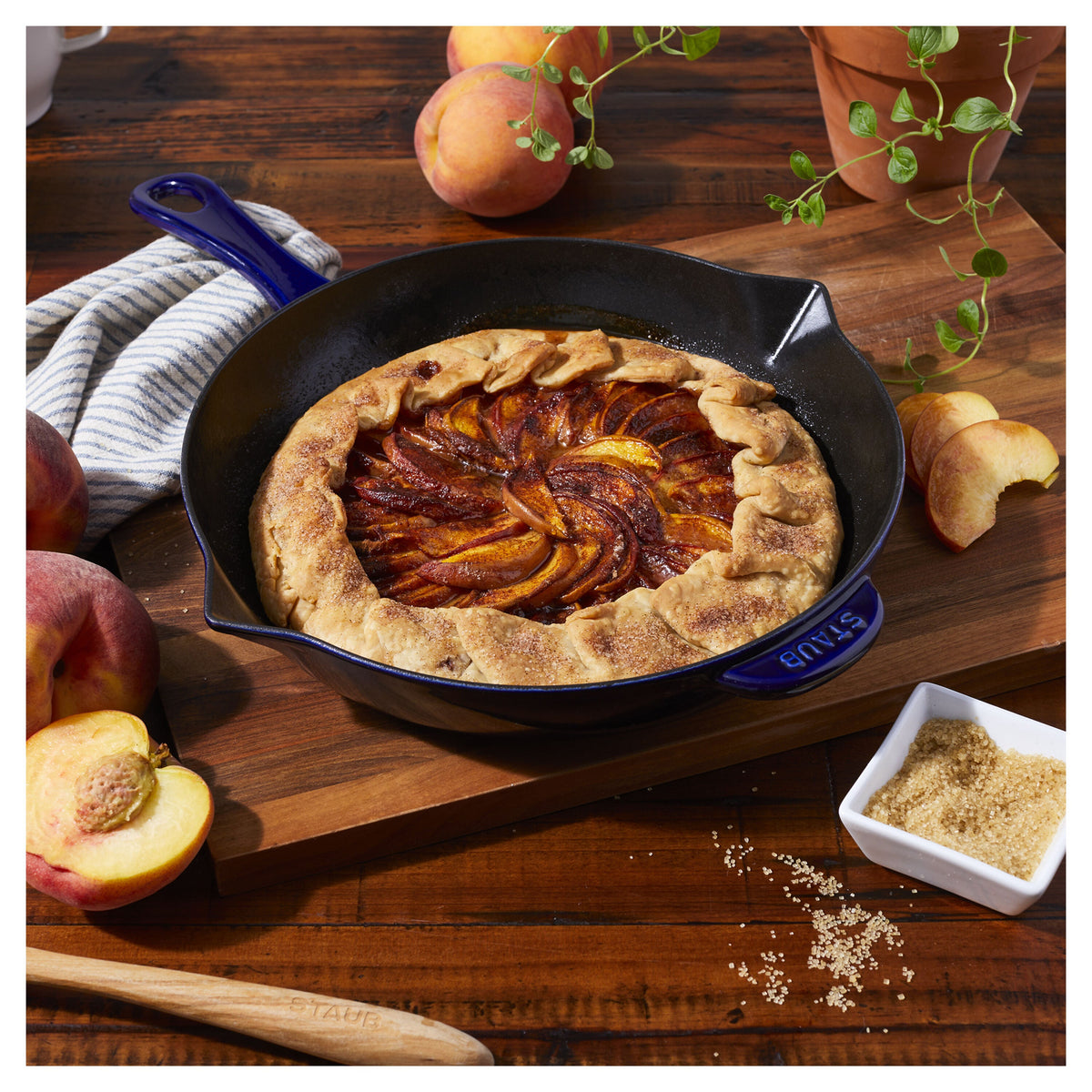 Peach galette in a cast iron skillet on a wooden table with peaches and a plant.