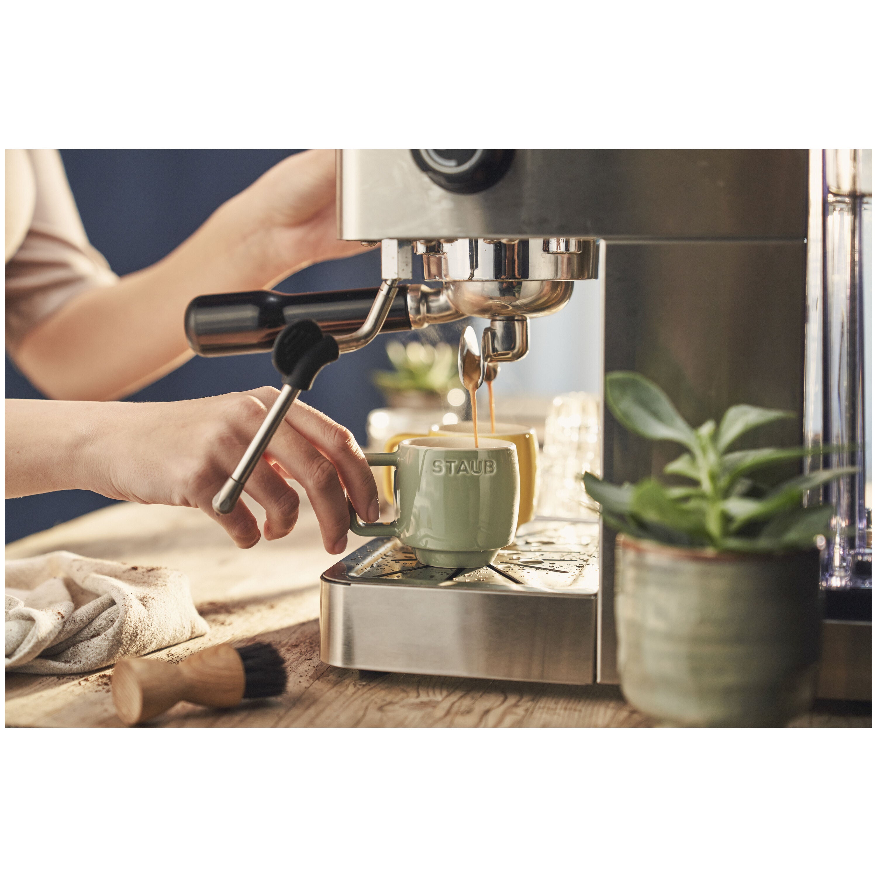 Person using an espresso machine to pour coffee into a Staub mug, with a plant in the background.