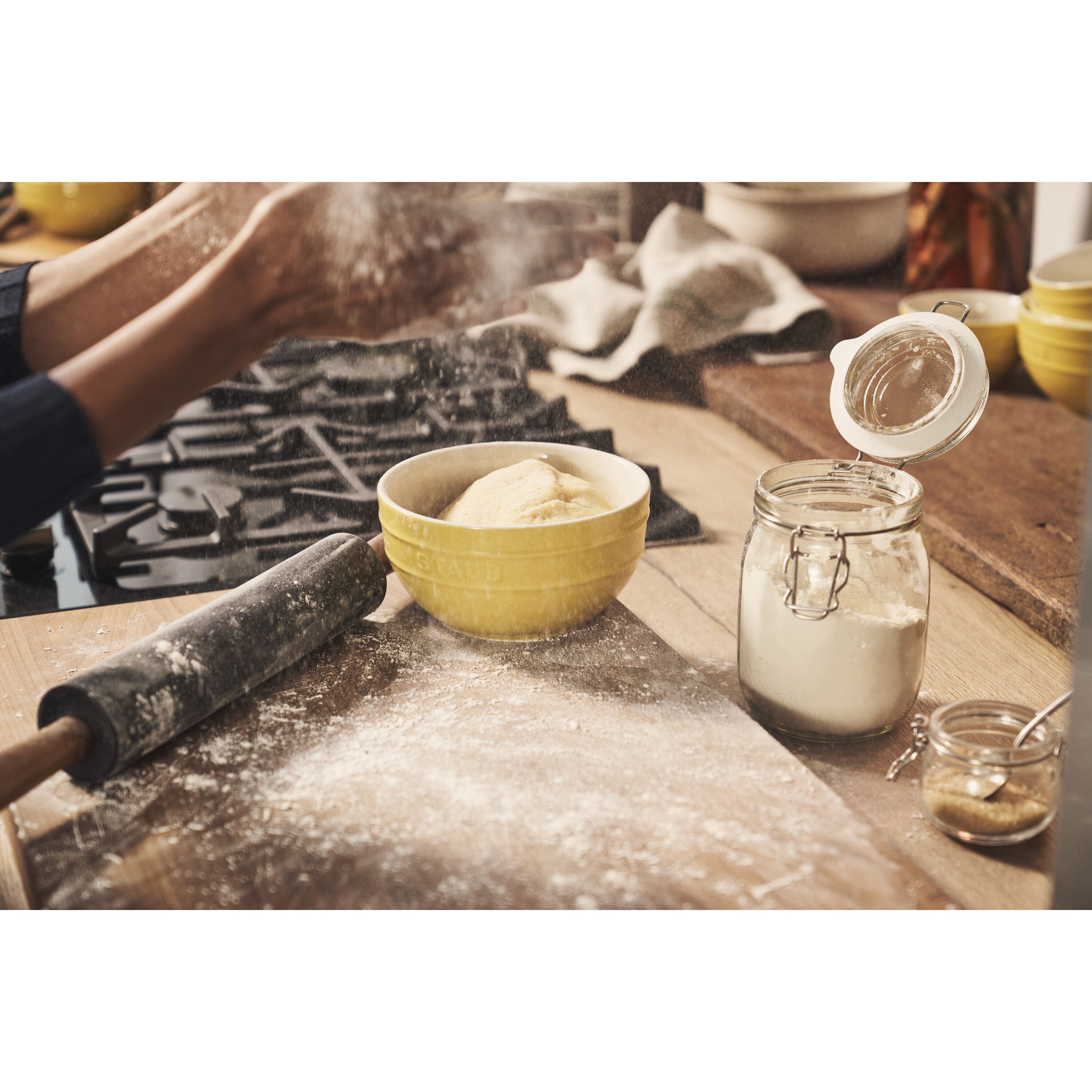 Person kneading dough with a rolling pin on a floured surface, surrounded by kitchen items.