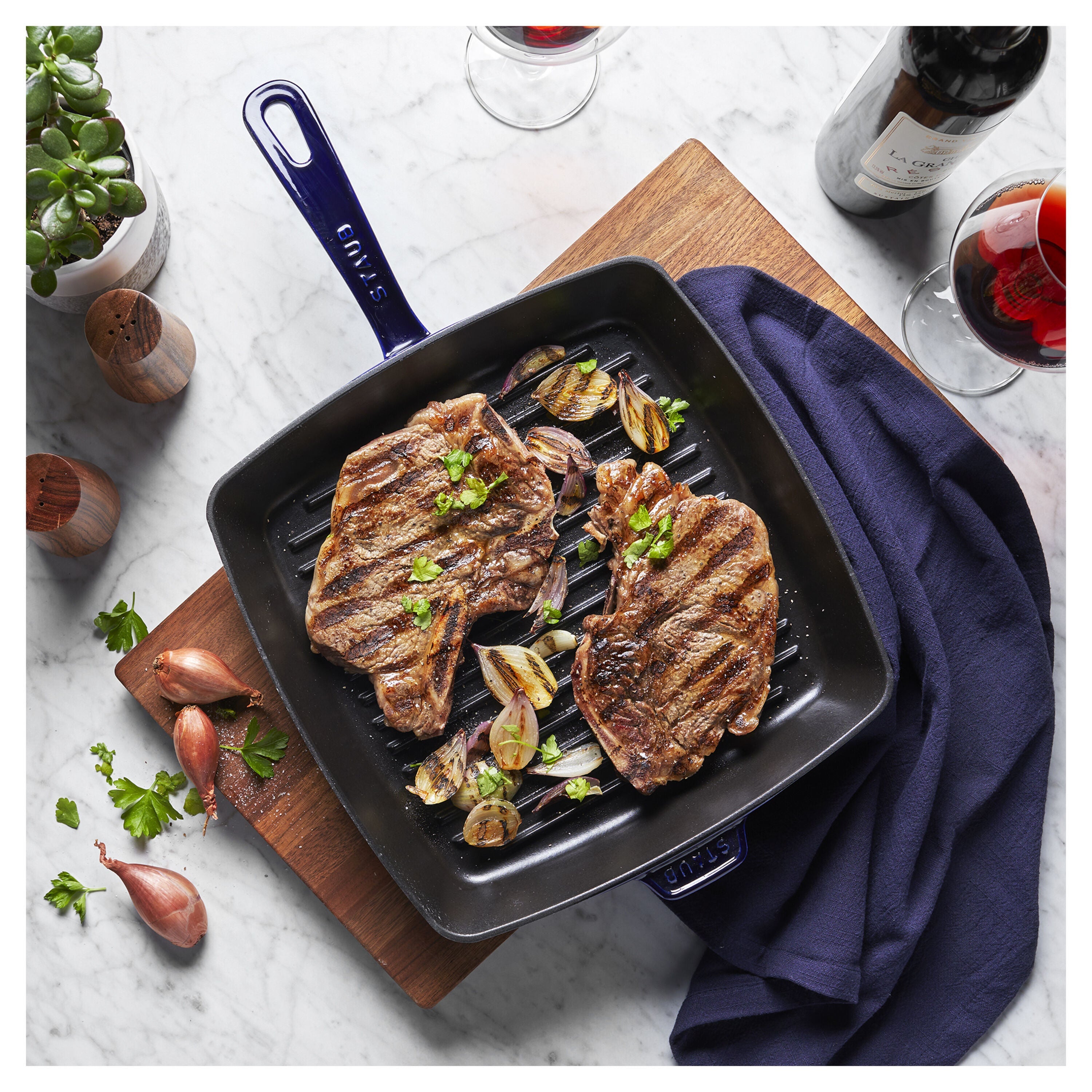 Grilled steak with garlic and herbs in a black grill pan on a wooden board, with a bottle of wine and glasses in the background.