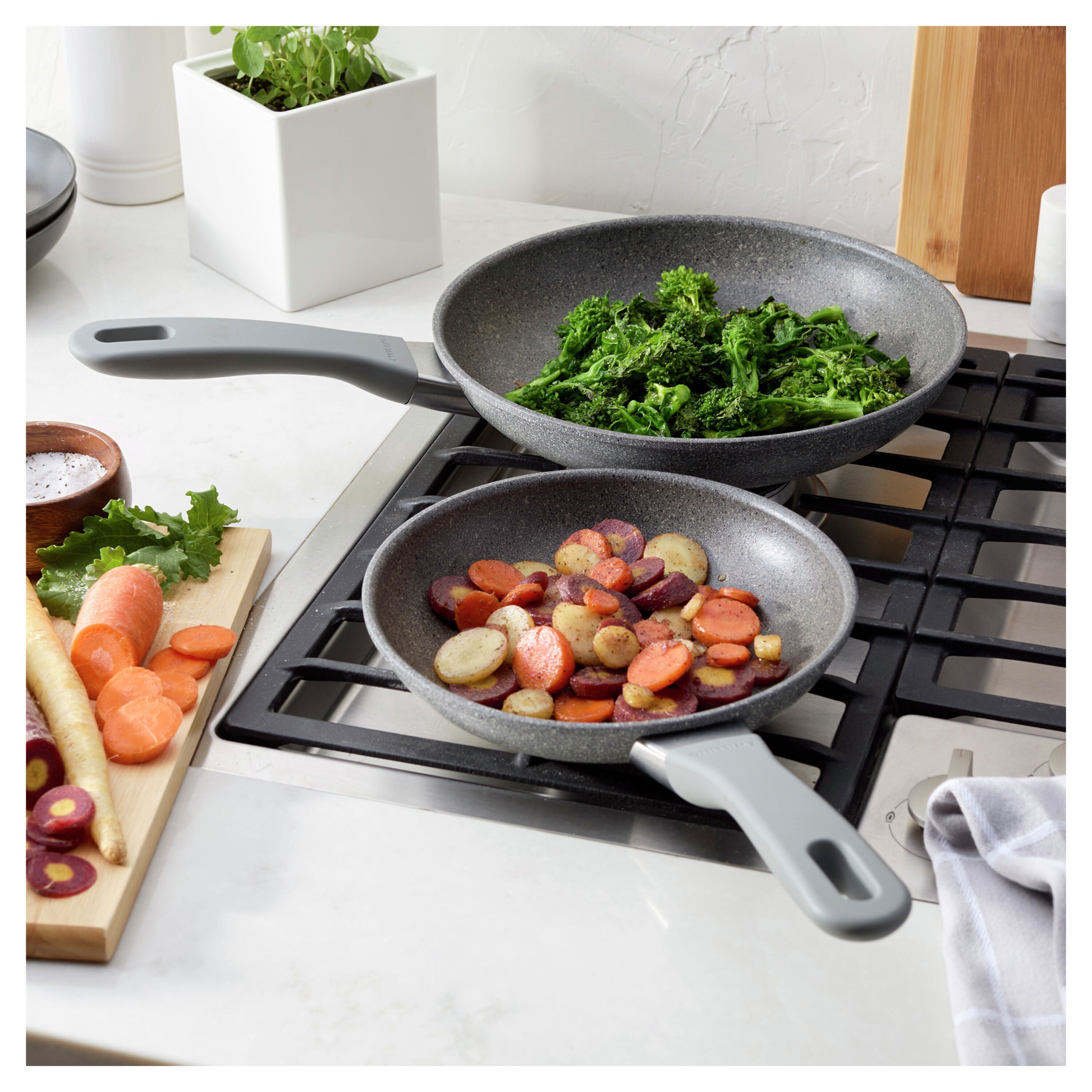 Two frying pans on a stove with vegetables inside, surrounded by kitchen items.
