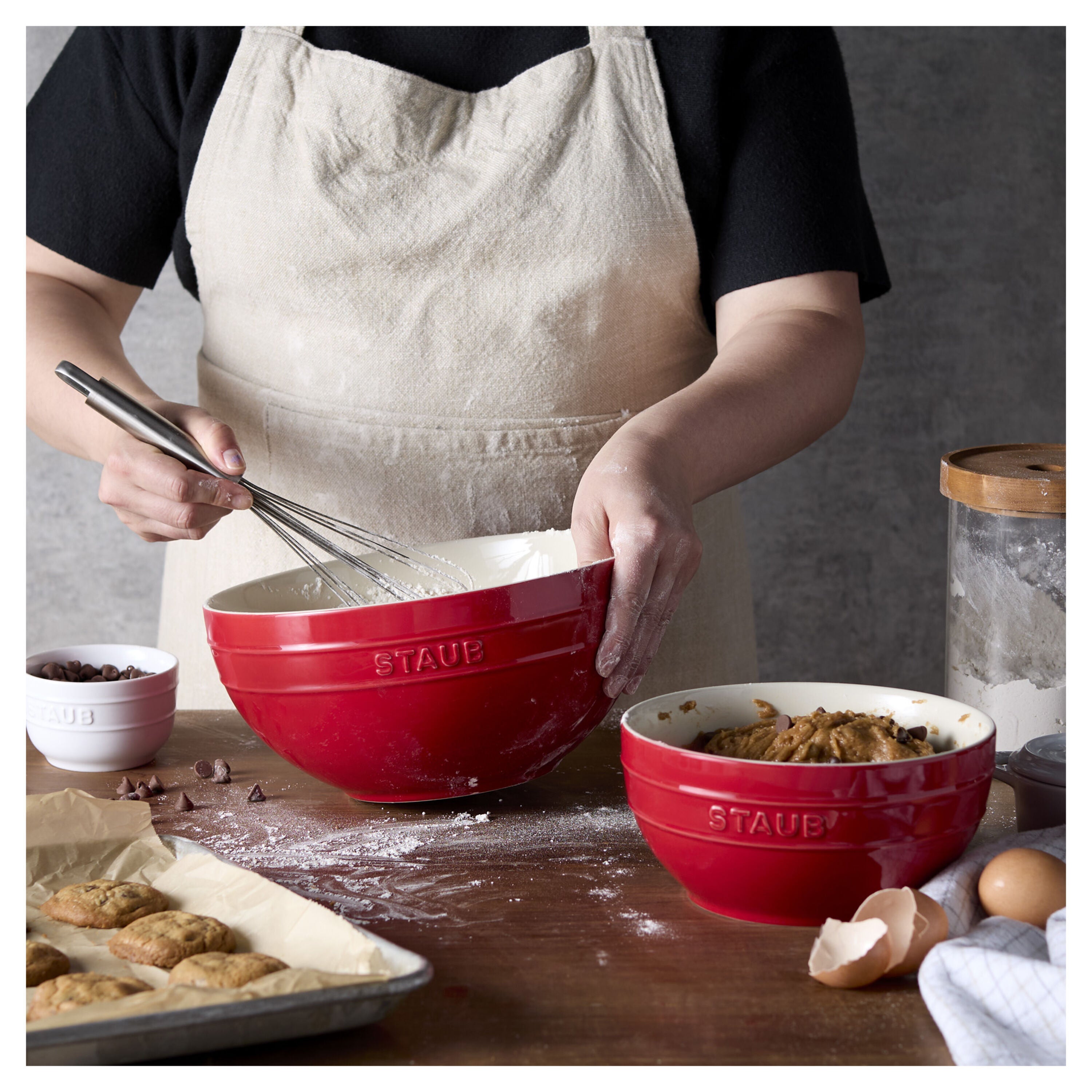 Person in a kitchen with red Staub bowls and baking ingredients.