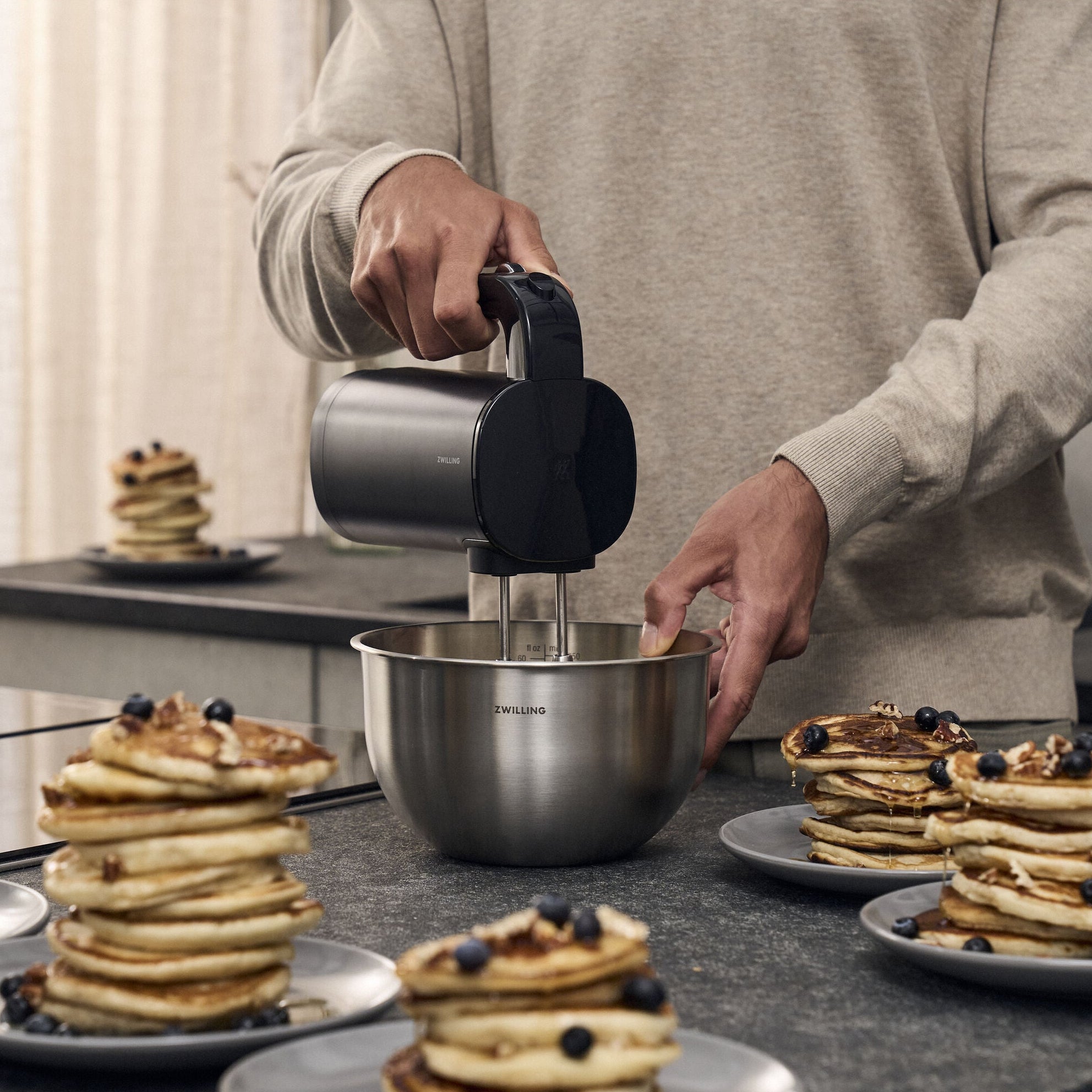 ZWILLING XTEND Cordless Hand Mixer in use, mixing batter in a bowl, surrounded by plates of pancakes on a kitchen counter.