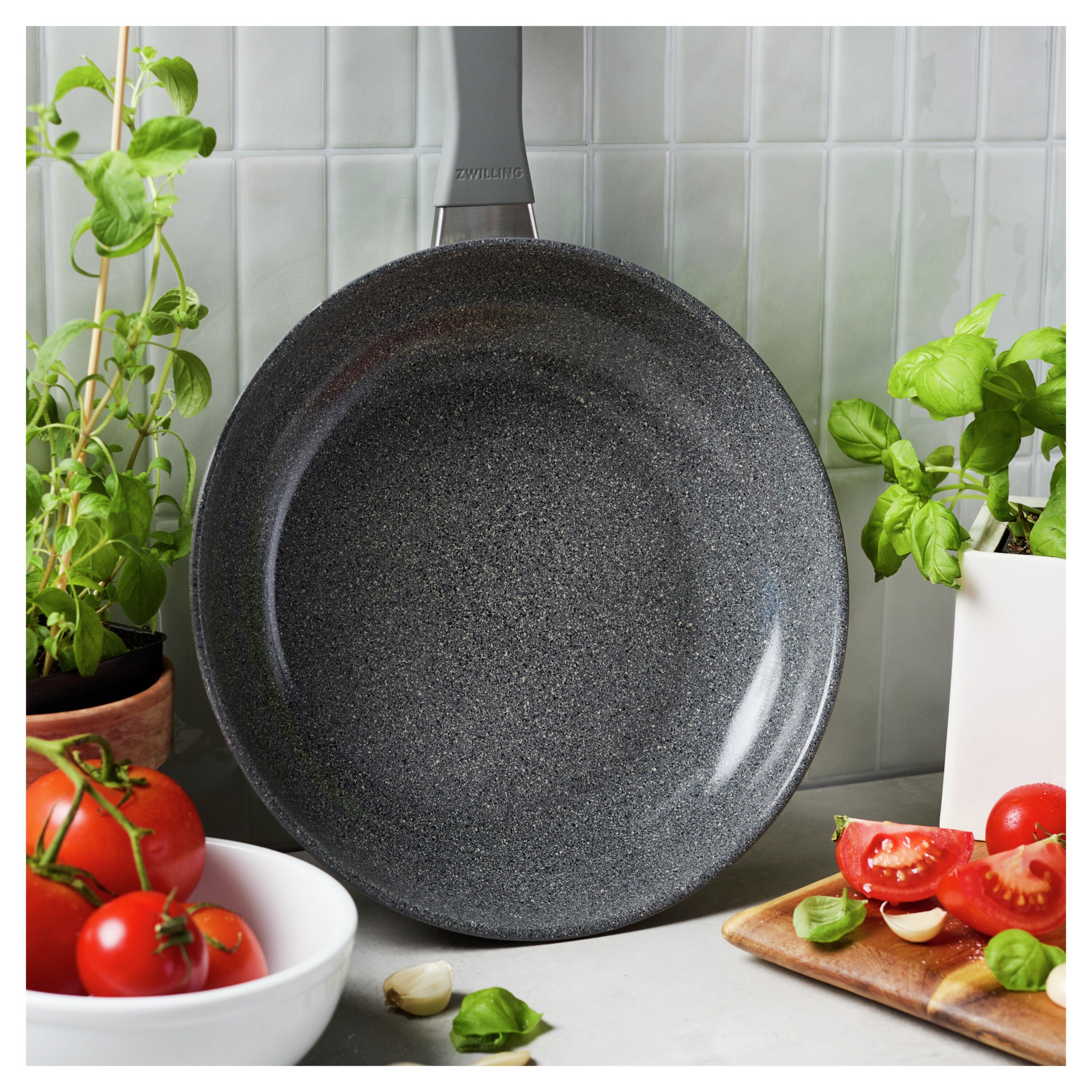 Black speckled frying pan on a kitchen counter with tomatoes and herbs.