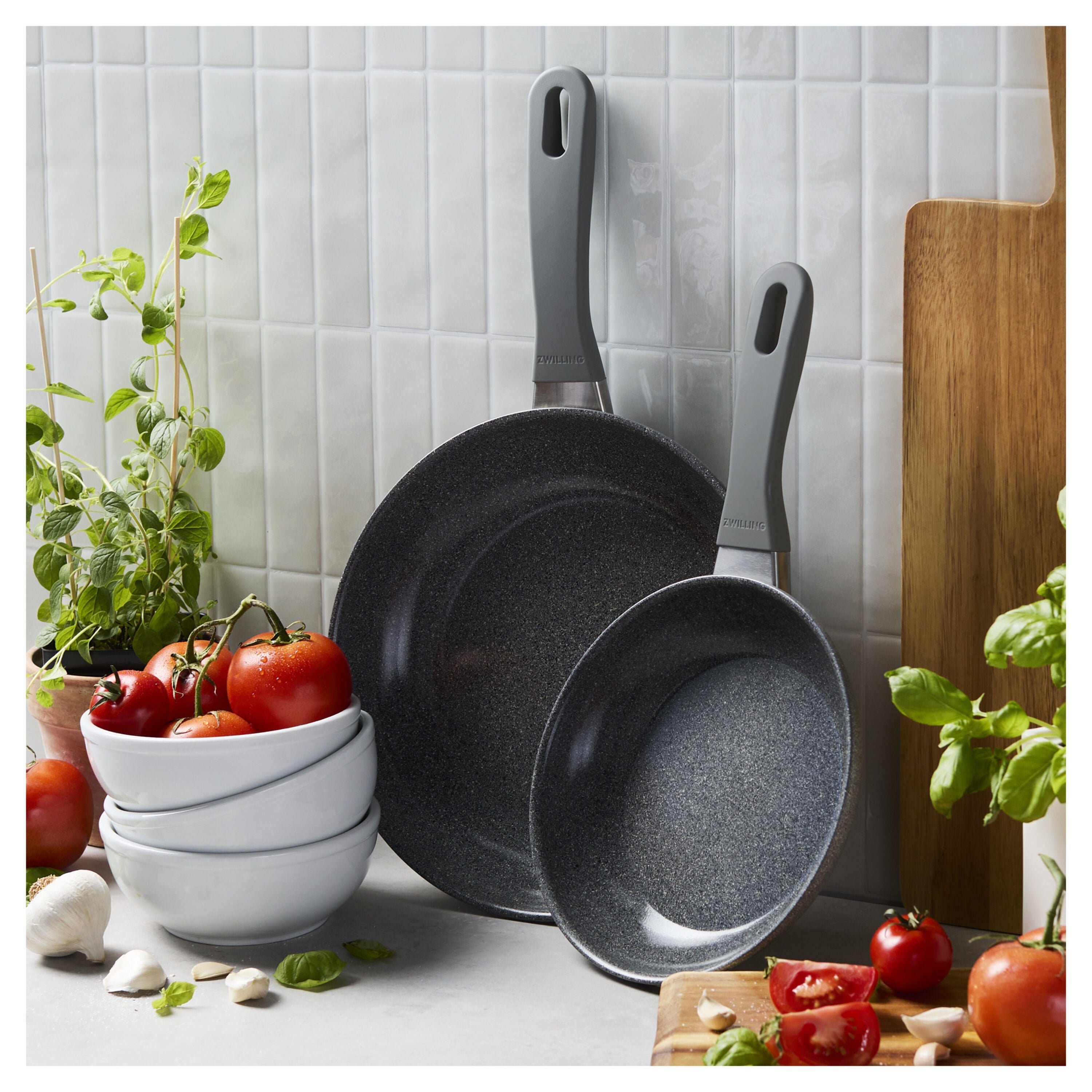 Set of gray frying pans on a kitchen counter with tomatoes and herbs.