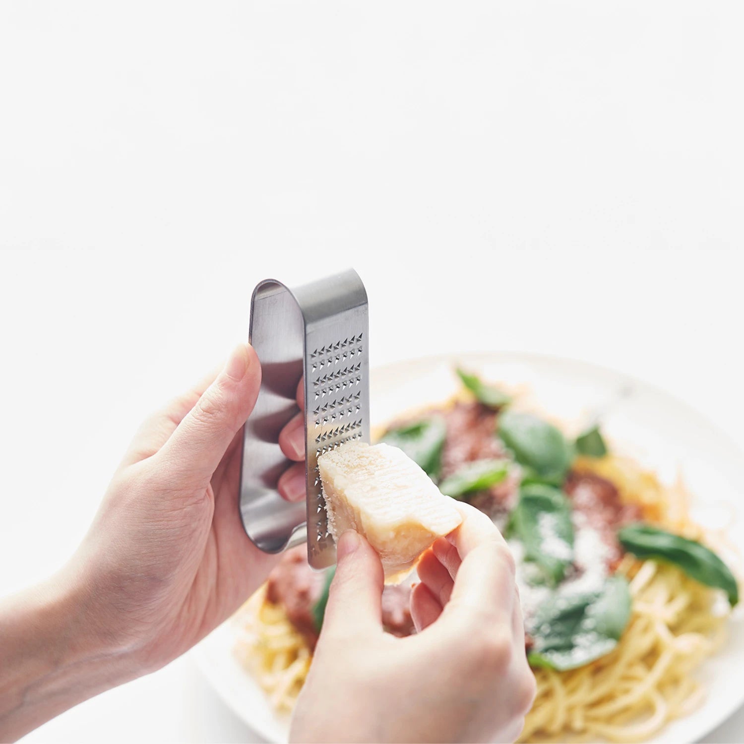 Hand holding a small metal grater over a plate of pasta with greens and cheese.