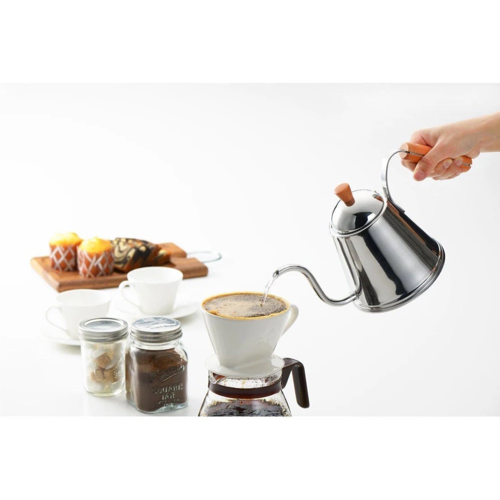 Person pouring coffee from a stainless steel pour-over coffee maker into a white cup on a white background.