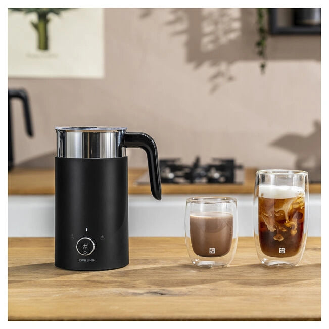 Black milk frother on a wooden surface with two glasses of coffee.