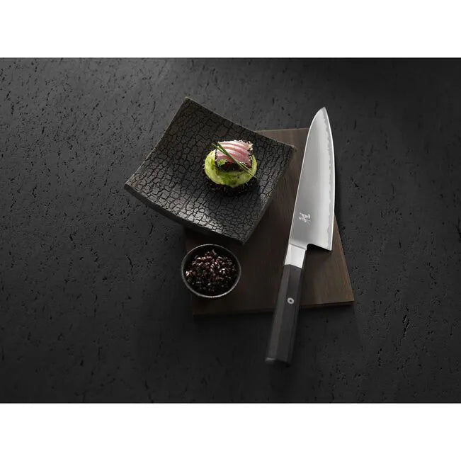 Chef's knife on a dark surface with a small dish of food and spices.