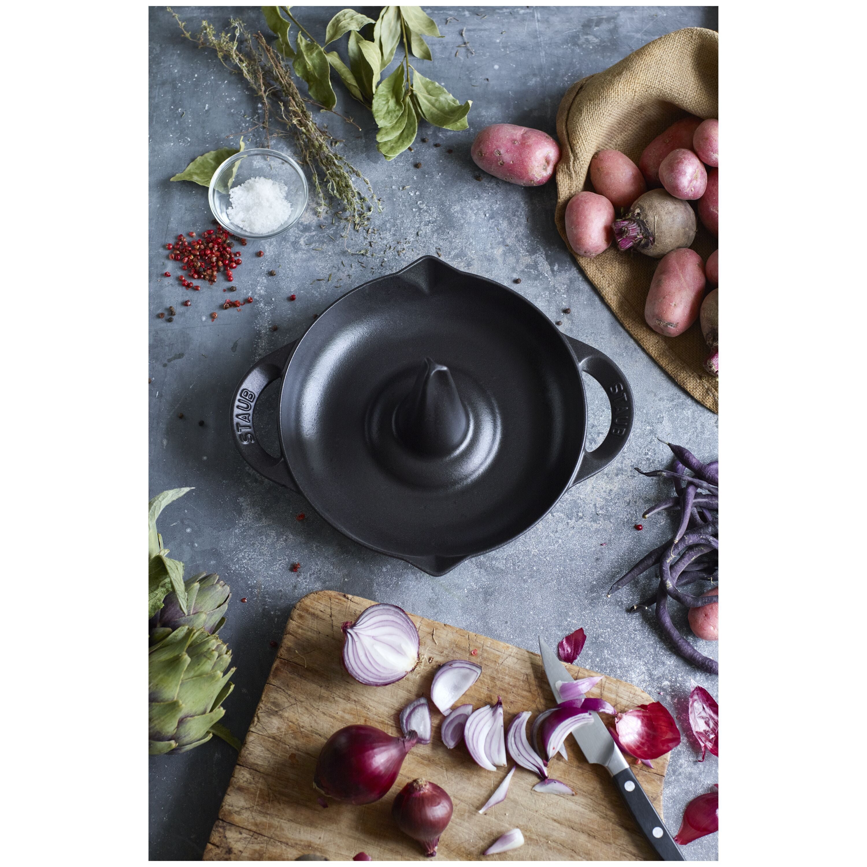Top view of STAUB Specialities Cast Iron Round Roaster 24 cm in black, surrounded by fresh vegetables on a rustic kitchen counter.