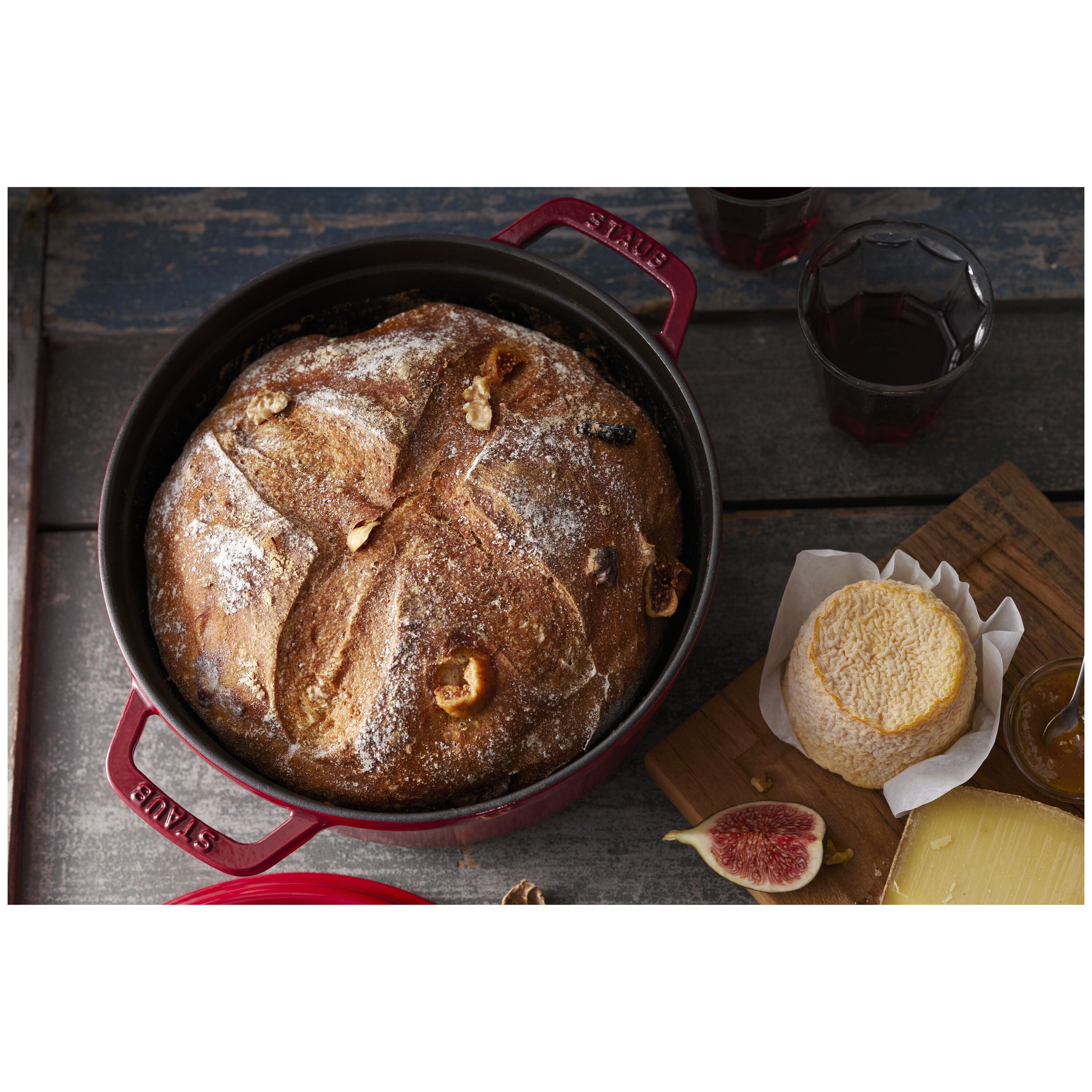 Baked loaf of bread in a red Staub cast iron skillet on a rustic wooden table.