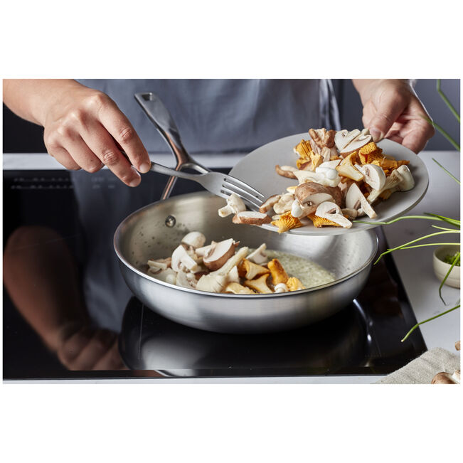 Person transferring mushrooms from a plate into a frying pan on a stove.