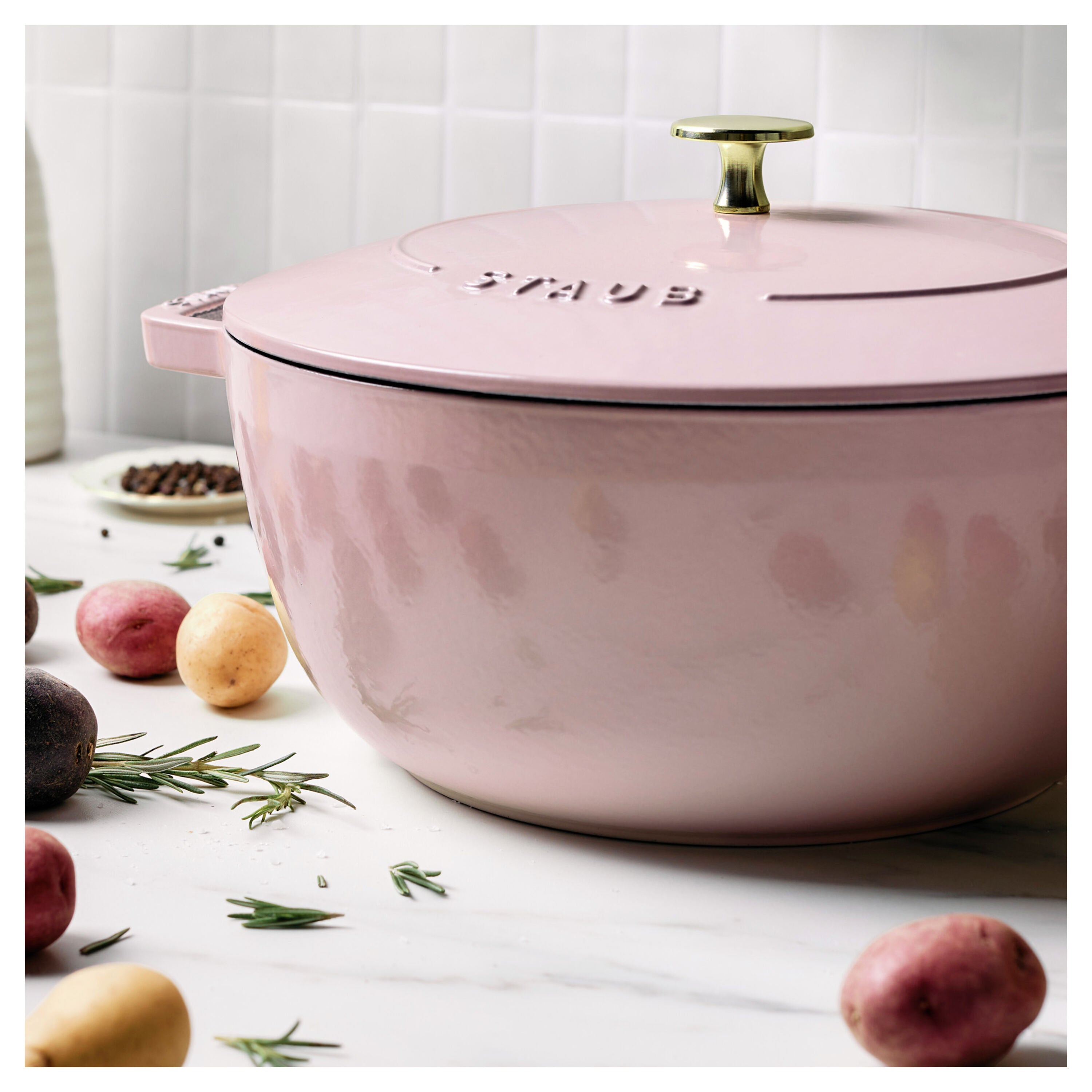 Pink Staub cookware pot on a white surface with potatoes and rosemary.
