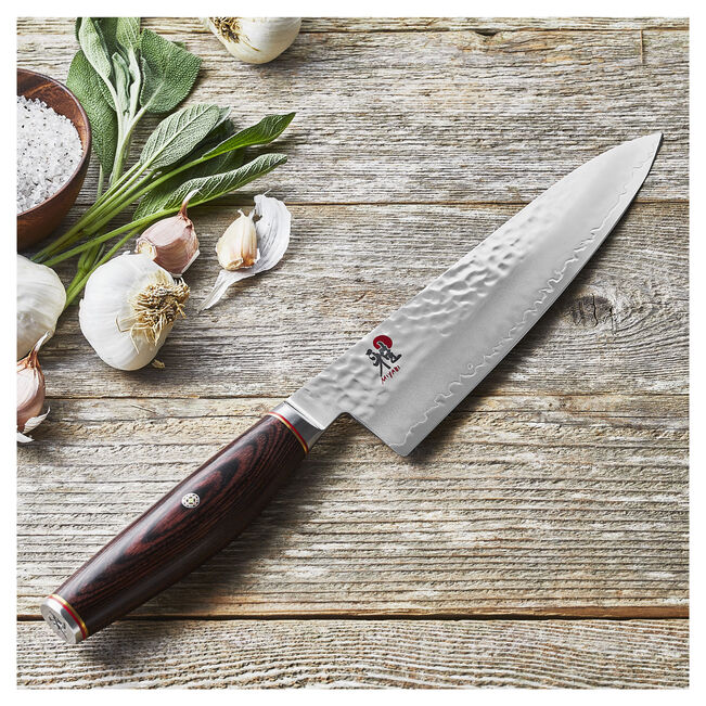 Miyabi 6000 MCT 8 Inch Gyutoh positioned on a wooden surface with fresh herbs and garlic in the background.