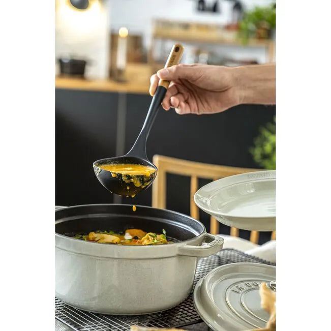Person ladling food from a pot into a pan with a kitchen setting in the background