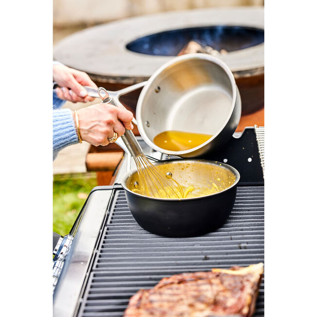 Person cooking outdoors, pouring a liquid into a pan with a whisk.