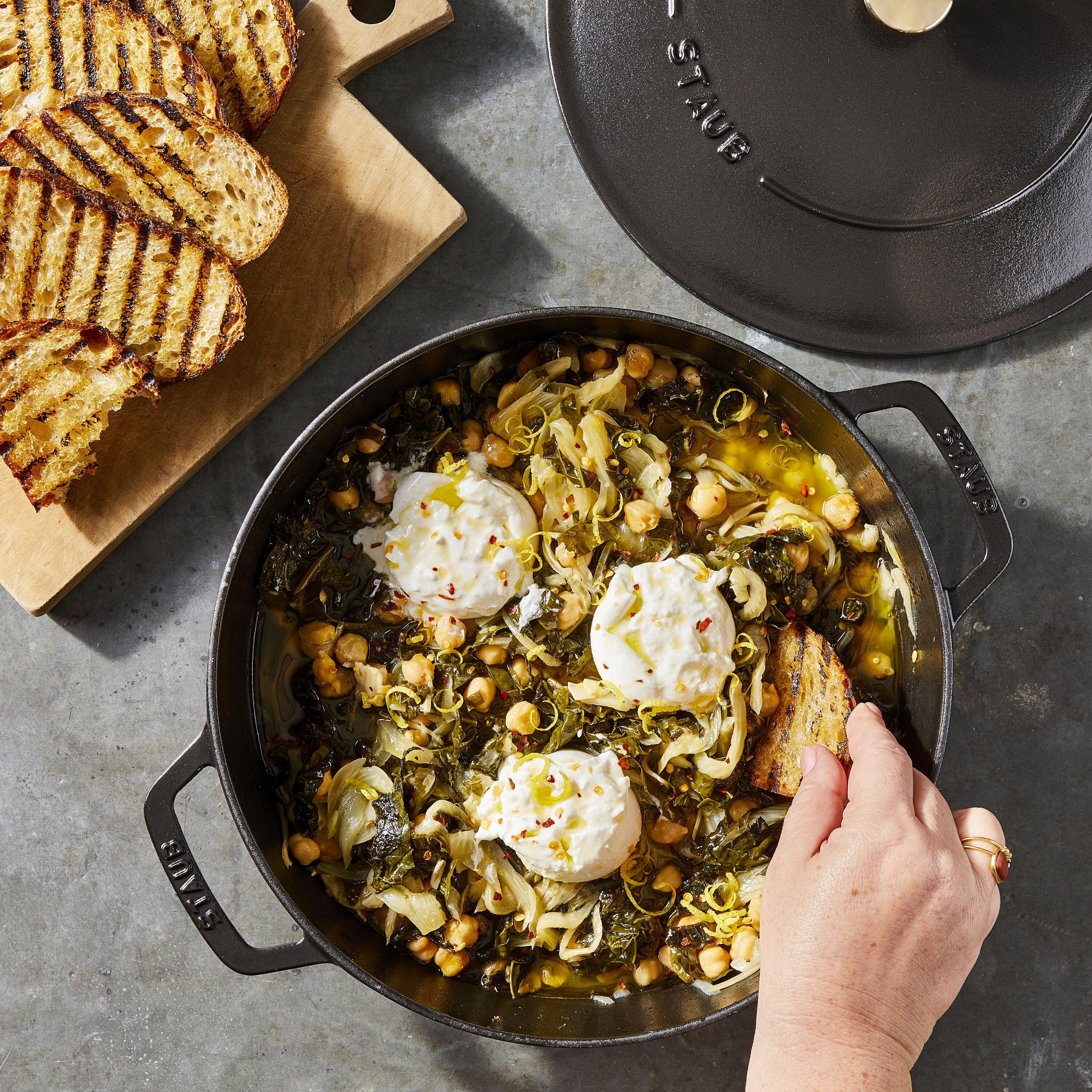 Dish of food with a hand holding a piece of bread, next to grilled bread on a cutting board.