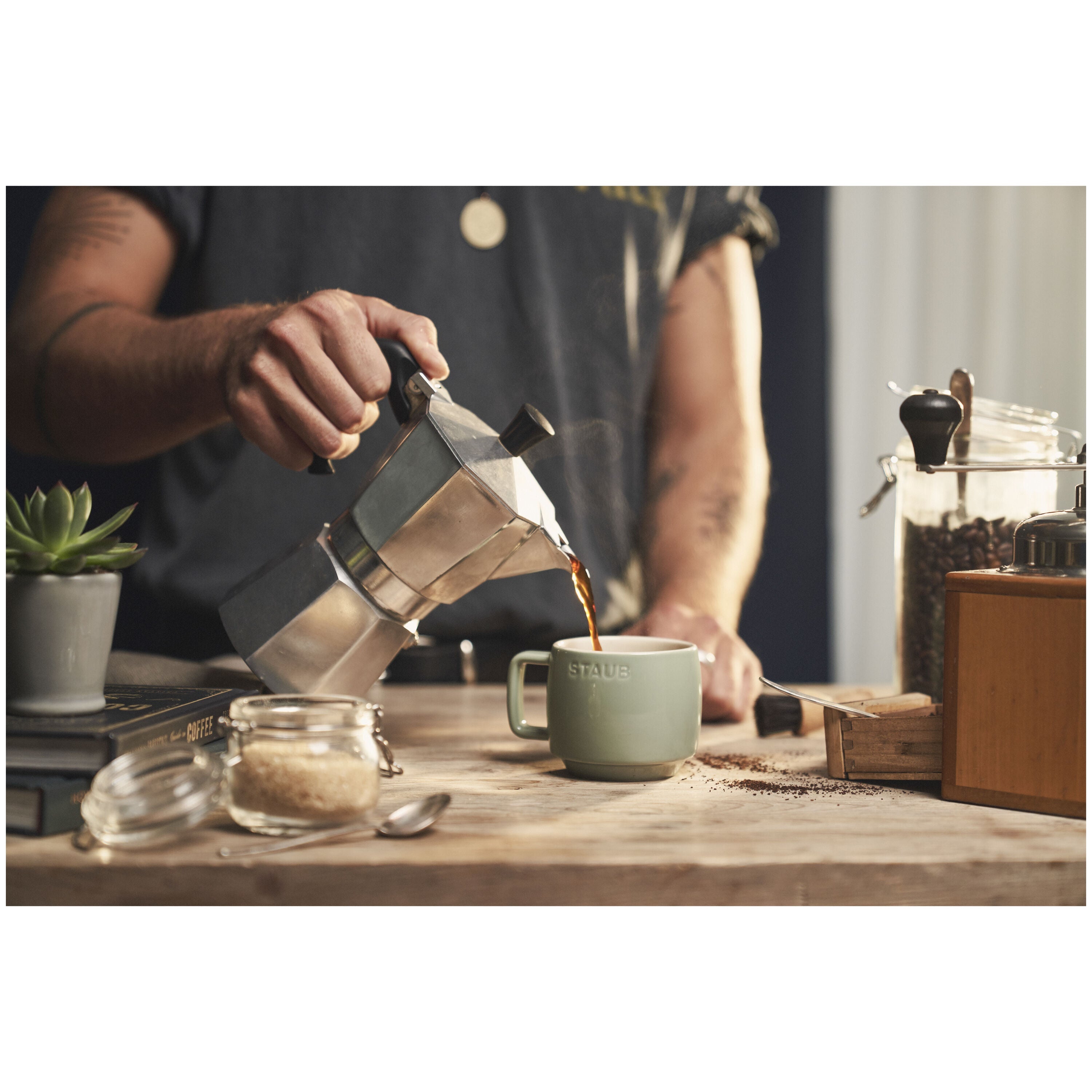 Person pouring coffee from a stovetop espresso maker into a green mug on a wooden table.