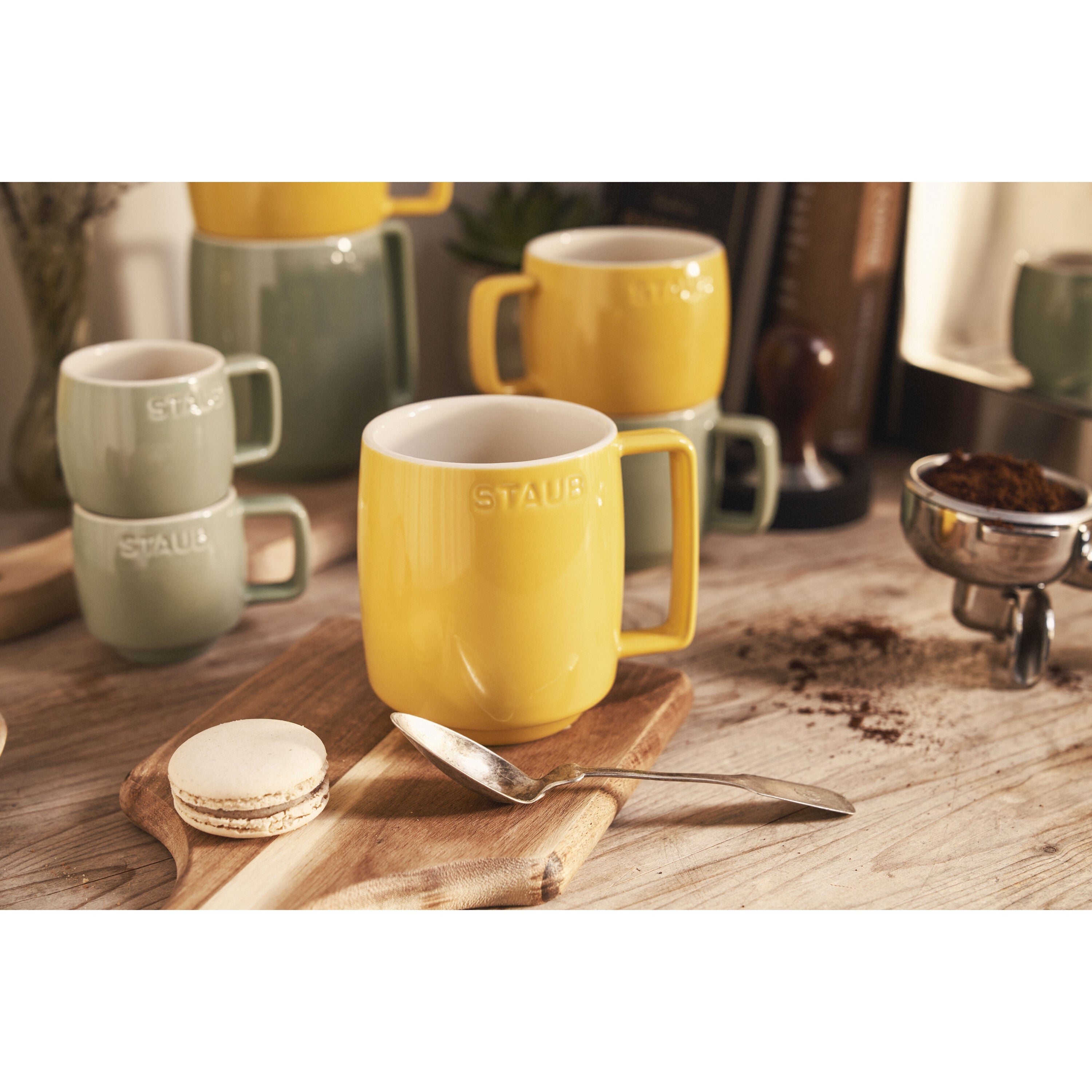 Yellow and green mugs on a wooden surface with a spoon and macaron.