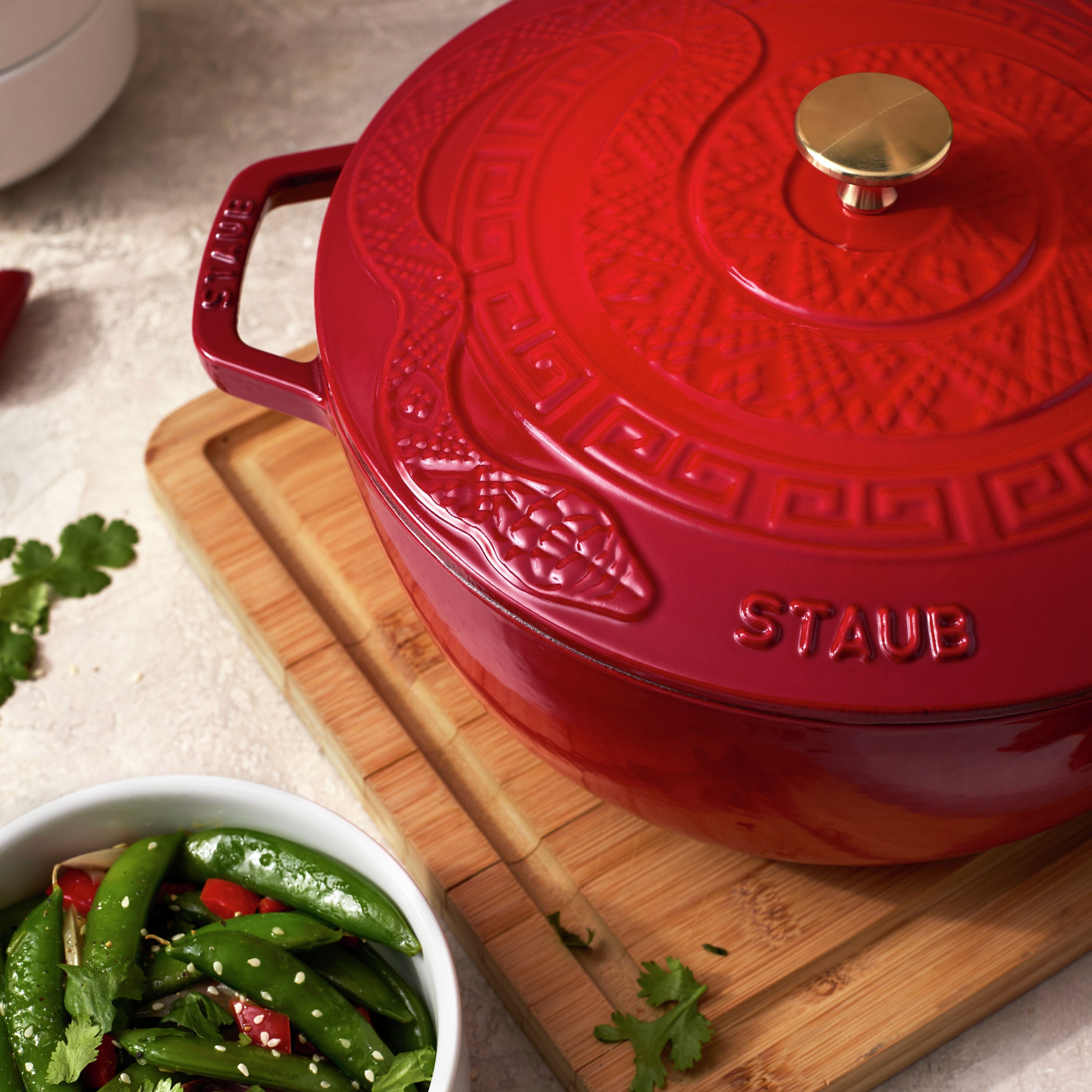 Red Staub cookware on a wooden cutting board with green vegetables.