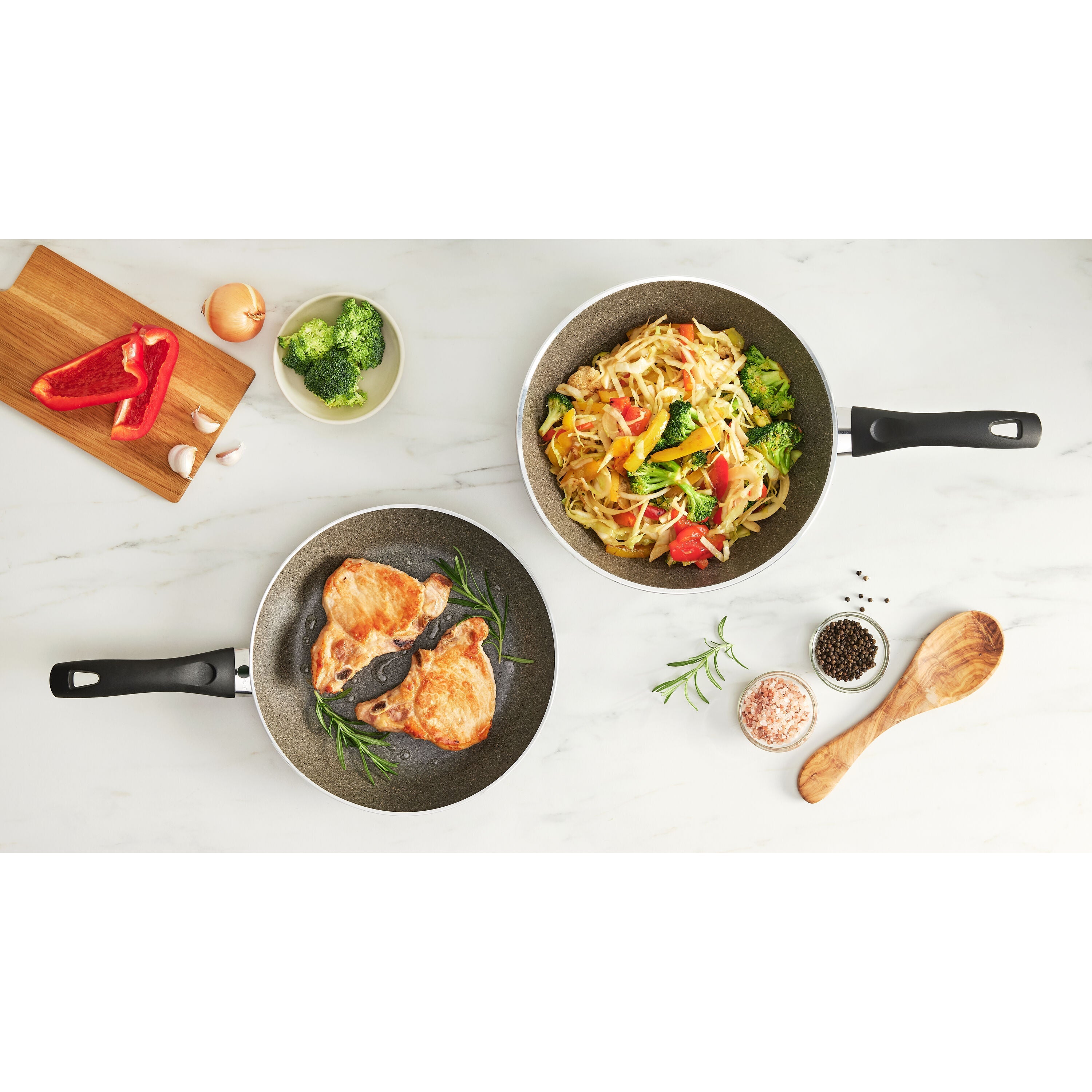 Two frying pans with food on a white surface, including broccoli and chicken.