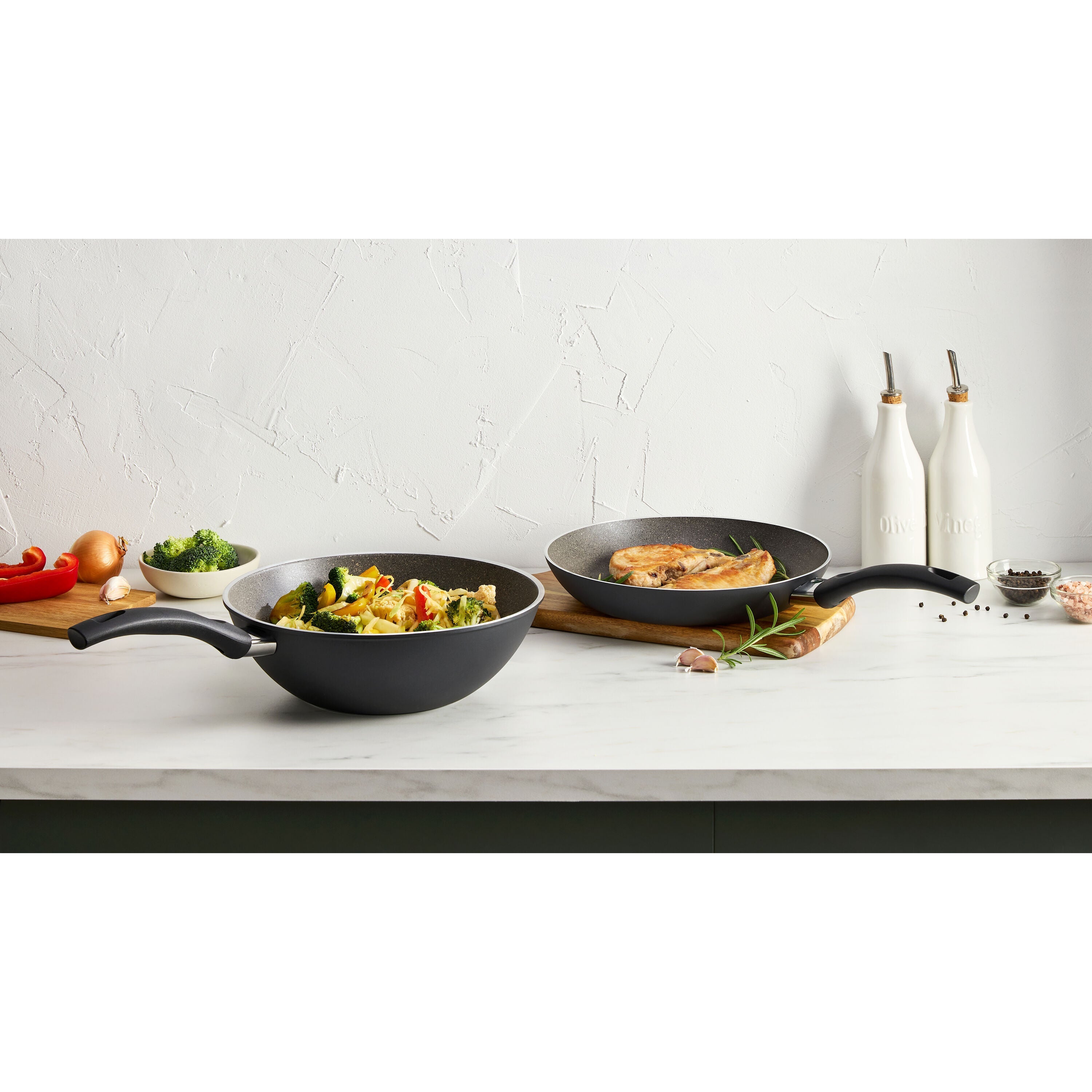 Two frying pans on a kitchen counter with food and ingredients.