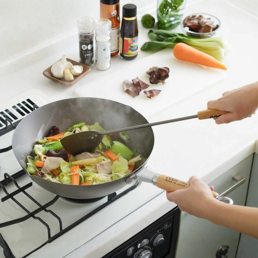 Person cooking vegetables in a wok on a stove with various ingredients around on YOSHIKAWA COOK-PAL Titanium Wok 